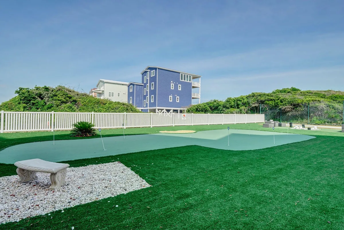 A picturesque exterior scene with green artificial turf, a dedicated golf putting area, a stone bench, and a charming blue house in the background. The area is designed for outdoor leisure, golf practice, and small gatherings.