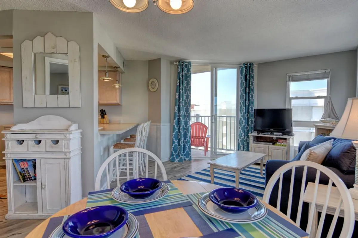 A bright, open interior with a dining table set with blue plates, a cozy living area with a blue sofa, and a red chair on the balcony offering views to the outdoors. The space is designed for casual family dining and relaxed gatherings, with easy access to a scenic balcony.