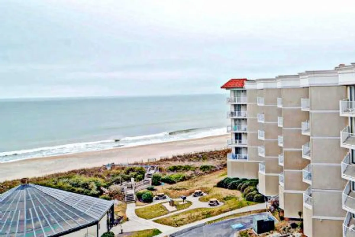 An exterior shot capturing the ocean view, pristine beach area, the building structure, and a landscaped garden. The scene embodies tranquility while showcasing the direct relationship between the balcony's vantage point and the surrounding environment.