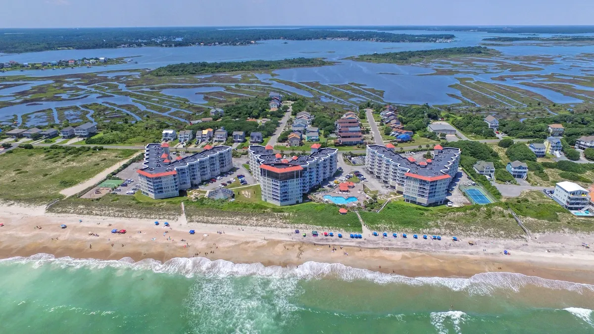 This exterior image features beachfront condos with a sparkling swimming pool, beach umbrellas, and the Atlantic Ocean waves just beyond. The scene captures a classic resort atmosphere, with inviting lounge areas and direct access to both the pool and beach.