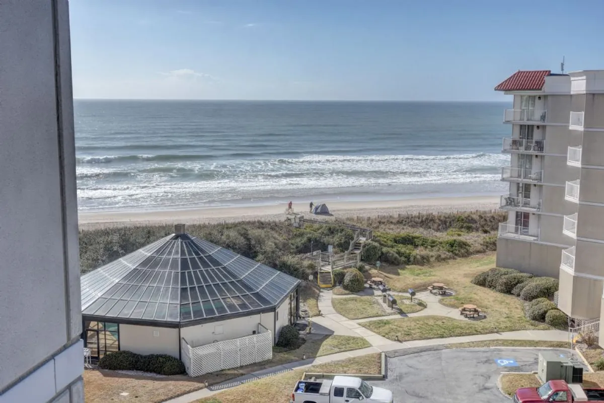 The image shows a serene beachfront scene at North Topsail Beach, North Carolina. The foreground features a charming picnic area and a covered pavilion overlooking the expansive sandy beach and gentle ocean waves. The scene exudes tranquility, with soft sunset lighting casting a warm glow, inviting guests to unwind, gather, or stroll along the shore.