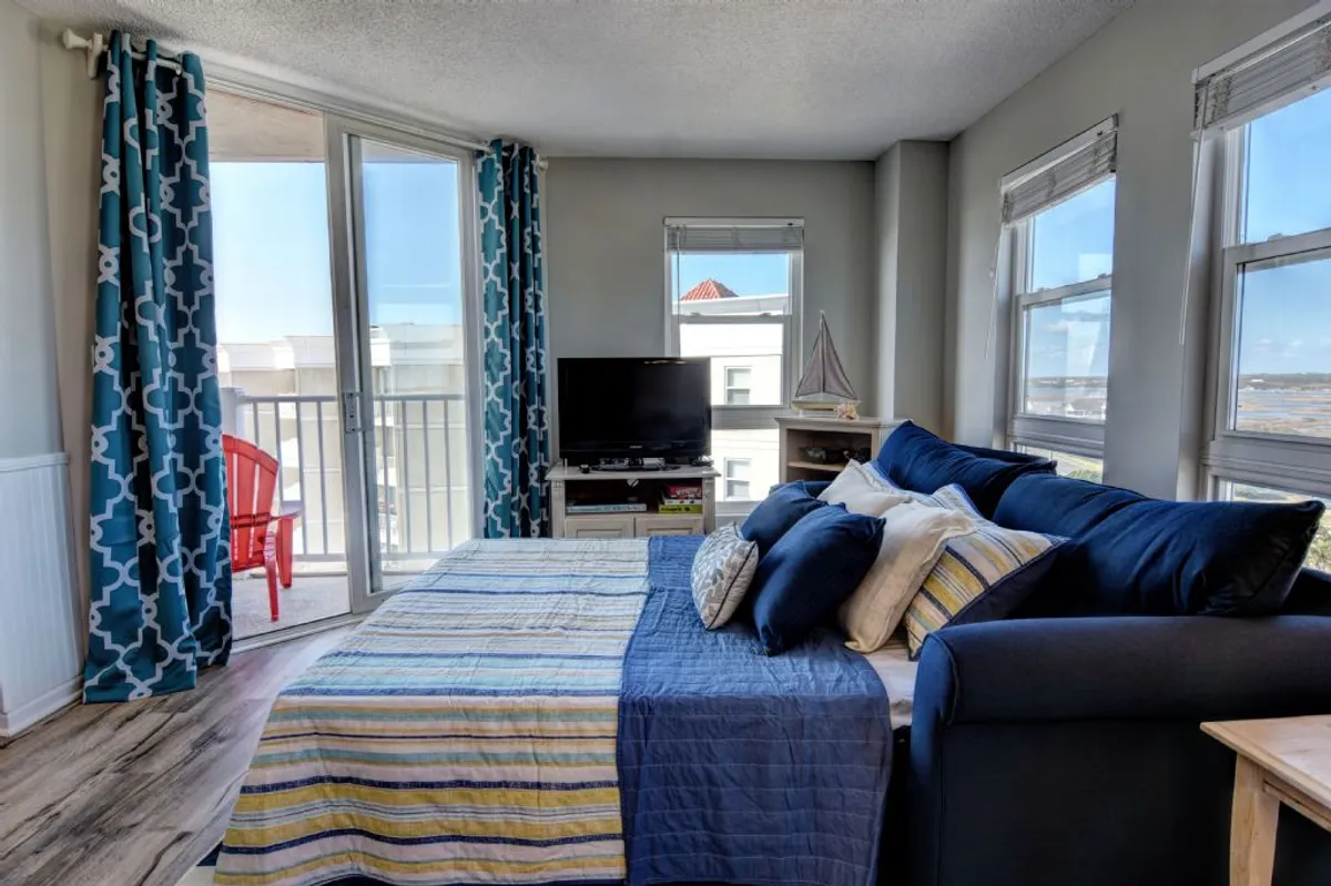 A bright, cozy bedroom with colorful bedding and a red chair placed on the balcony outside. Natural light pours in through large windows, offering a glimpse of the inviting balcony space and the atmosphere it provides.
