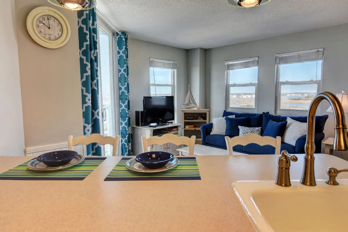 The image displays a bright kitchen counter with plates set for a meal, a blue sofa in the background, and large windows offering natural light and potential ocean views. The layout suggests a blend of cooking, entertaining, and enjoying the scenery.