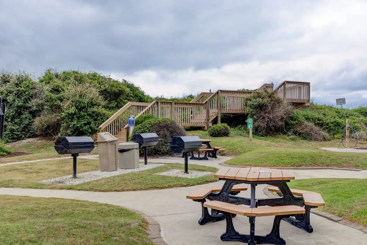 An inviting outdoor communal area featuring picnic tables and barbecue grills set on a landscaped lawn, with a wooden staircase leading to nearby amenities. The relaxed setup encourages social gatherings and alfresco dining.