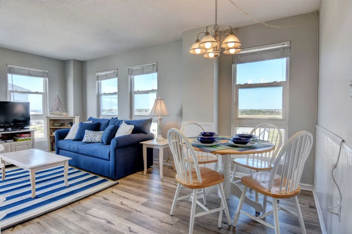 A bright, cheerful room with a blue sofa, dining table set for a meal, and large windows letting in sunlight. The fresh tile flooring and uplifting decor create a warm, welcoming atmosphere, ideal for shared moments and relaxation.