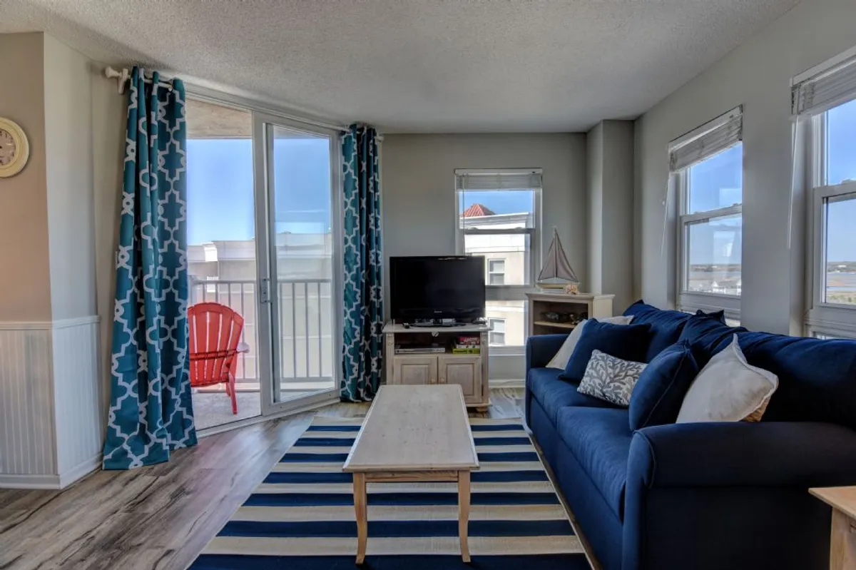 This image features a spacious living room with a blue sofa, striped rug, red accent chair, large windows, and a wall-mounted TV. The room is filled with natural light and feels airy yet cozy, with a relaxed, beach-inspired decor palette.