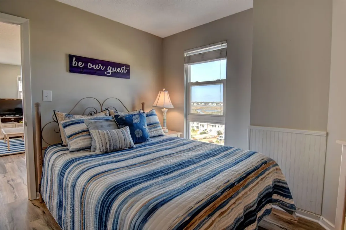 This image shows a cozy bedroom featuring a bed with striped bedding, a nightstand with a lamp, and a window that lets in natural light—creating a serene and welcoming atmosphere.