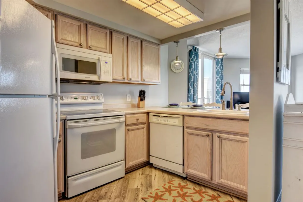This image features a warmly lit kitchen with white appliances, light wood cabinetry, a decorative orange rug, and a window with blue curtains. The setup suggests a fully equipped space ideal for family meals, baking, and entertaining guests.