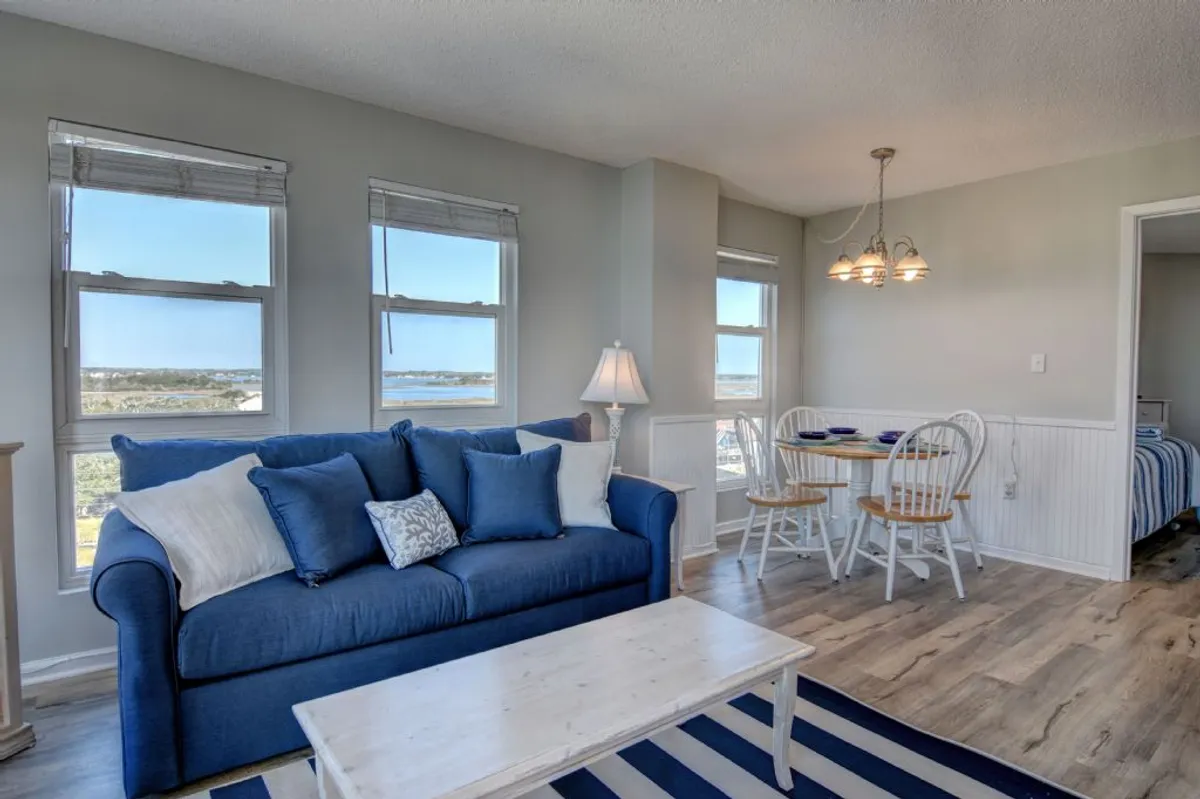 A bright living and dining area featuring a comfortable sofa, dining table, large windows, and a lamp. The room looks inviting for relaxation, socializing, or accommodating guests.