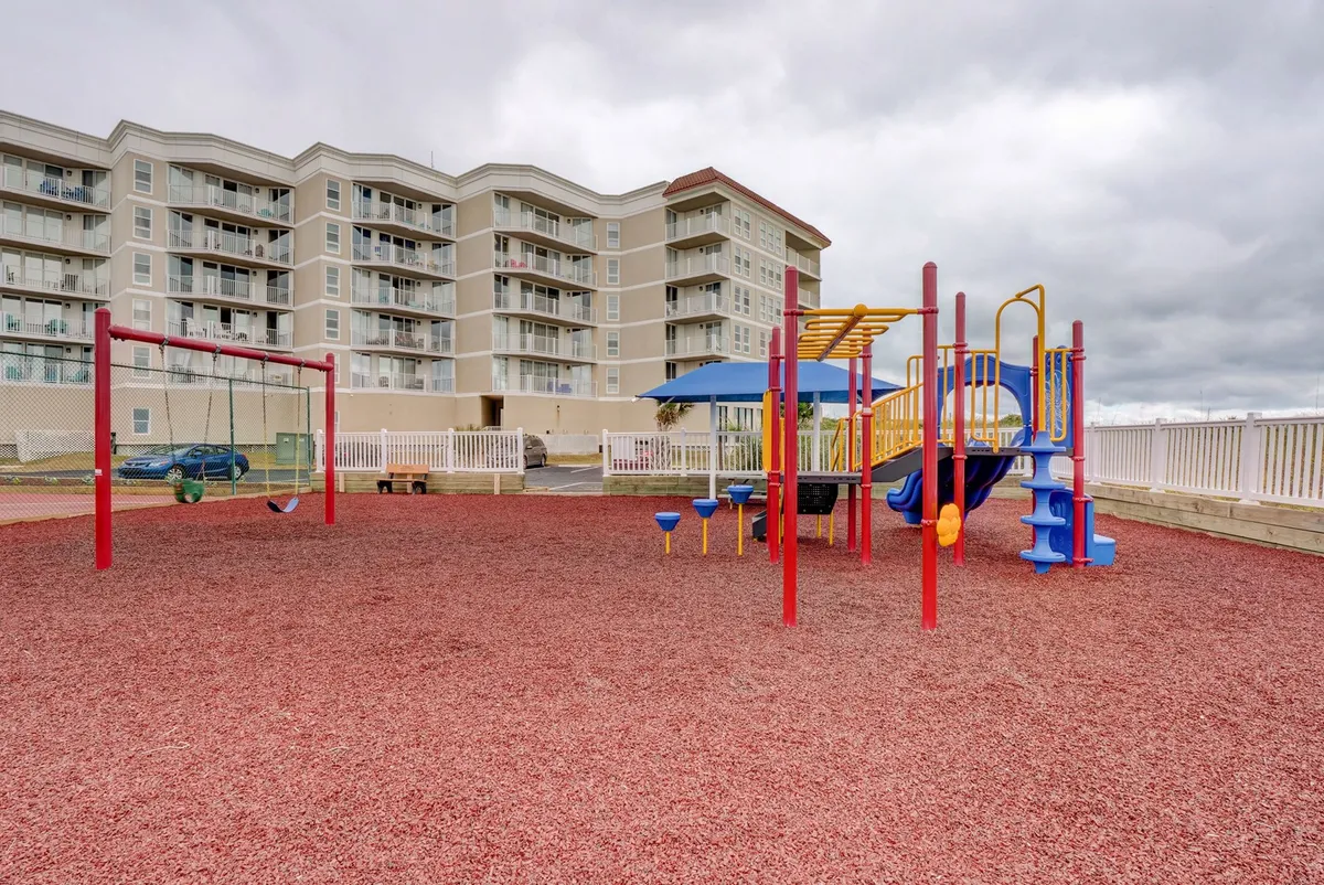 A vibrant, colorful playground with swings and play structures set on red rubber mulch, with a residential building in the background. Several children are seen playing and families gather nearby, creating an atmosphere of lively outdoor fun.